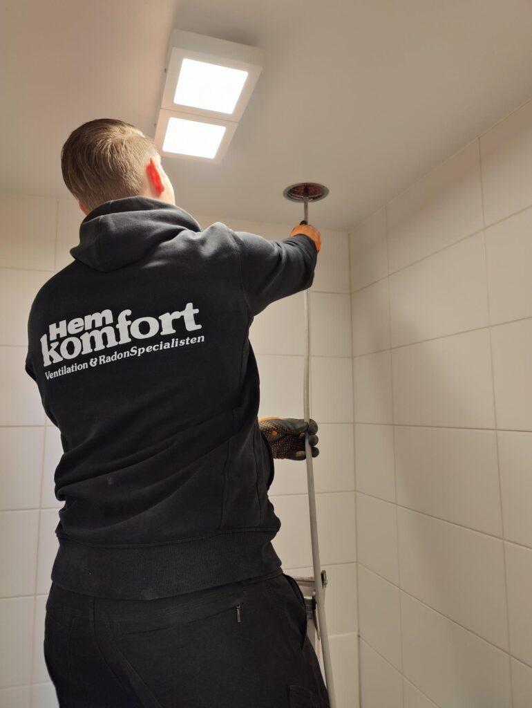 Hemkomfort-tekniker står på en pall och rengör ett ventilationsgaller i taket i ett vitt kaklat badrum. Personen bär en svart hoodie med Hemkomfort logotyp på ryggen.